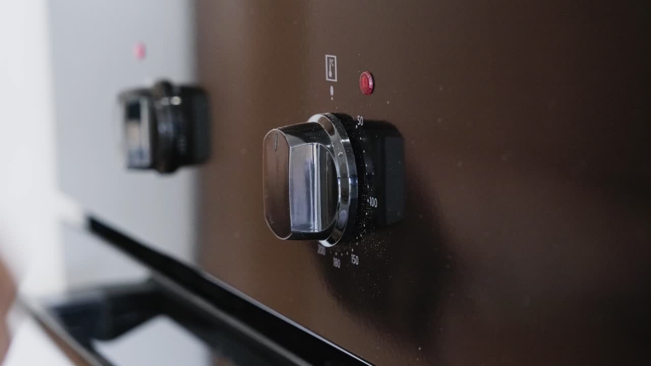 Hand operating dual oven control switches with red indicator lights. Close-up view of fingers turning knobs on modern appliance panel. Kitchen cooking preparation with clear tactile interaction