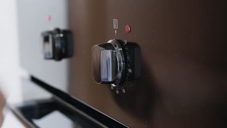 hand operating dual oven control switches with red indicator lights. close-up view of fingers turning knobs on modern appliance panel. kitchen cooking preparation with clear tactile interaction