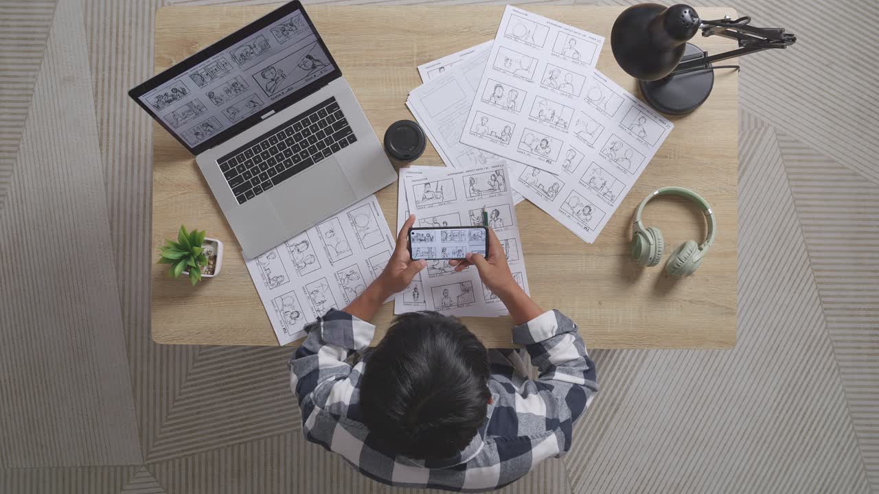 Top View Of Asian Male Artist Looking At Sketching Concept On Smartphone While Drawing Storyboard On The Table With A Laptop And Headphones In The Studio