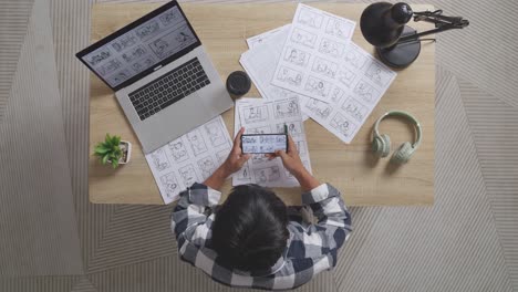 vista superior de un artista masculino asiático mirando un concepto de dibujo en un teléfono inteligente mientras dibuja un guión gráfico en la mesa con una computadora portátil y auriculares en el estudio