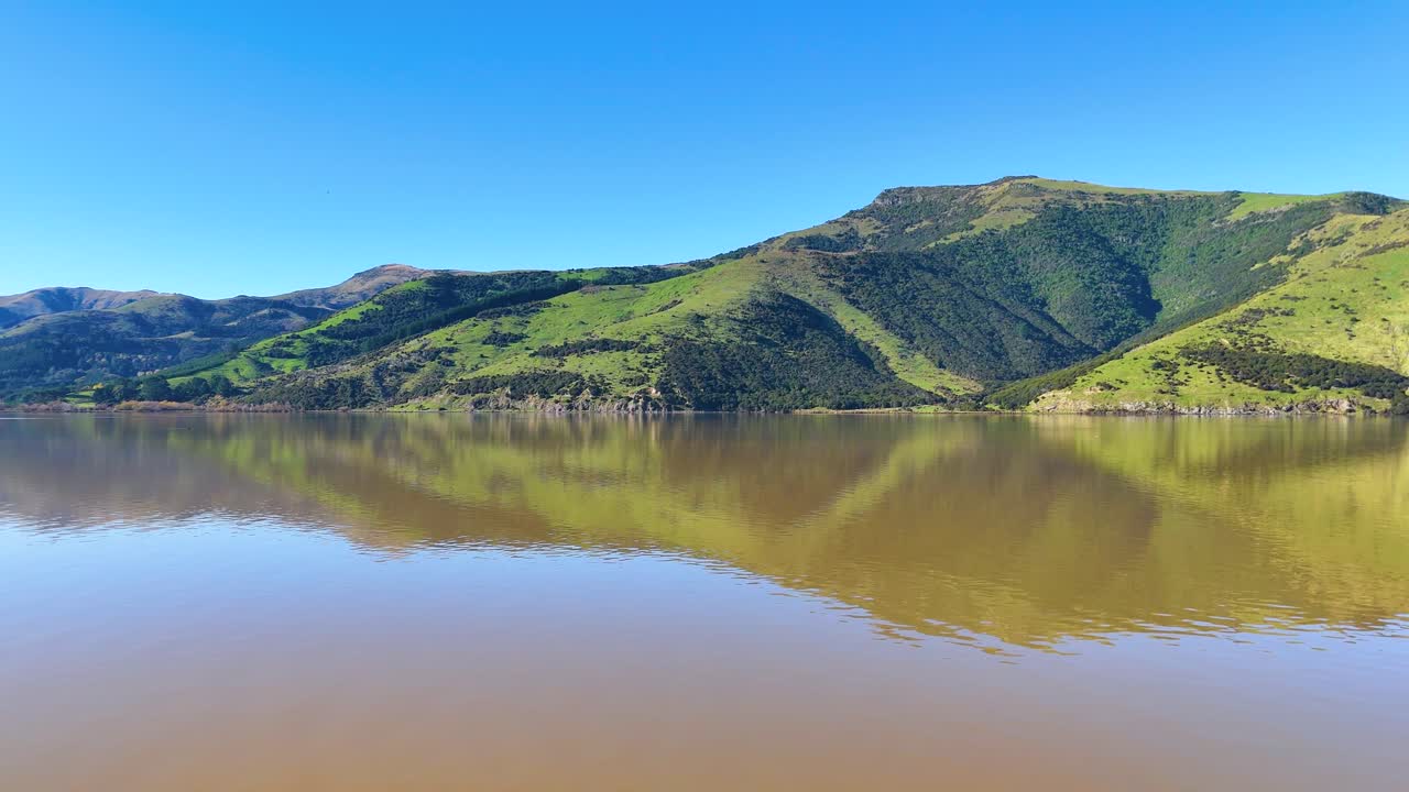 Calm lake reflecting lush green hills under clear blue skies in Akaroa, captured with smooth camera movement