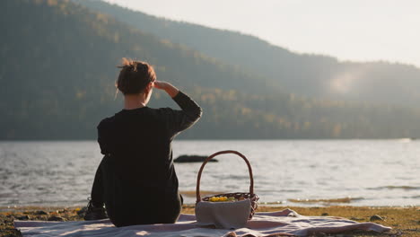 lady admires breathtaking view on picnic. relaxed woman looks at lake covering face against sunlight. solitude with nature sitting on blanket with basket