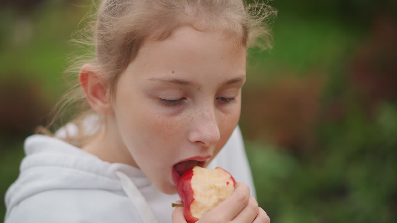 Young girl eating apple outdoors, hoodie worn by preteen with freckles takes slow bite of crisp red apple in lush garden, candid closeup captures thoughtful expression, summer daylight and soft bokeh