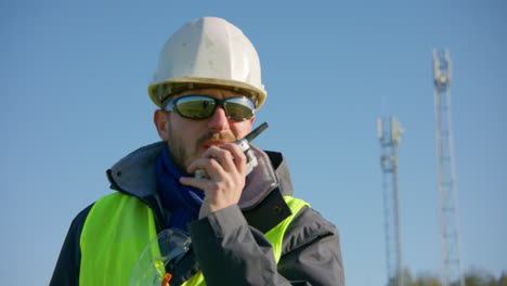 Male-engineer-speaking-over-the-walkie-talkie-with-cellular-towers-in-the-background-while-switching-subject-focus,-closeup