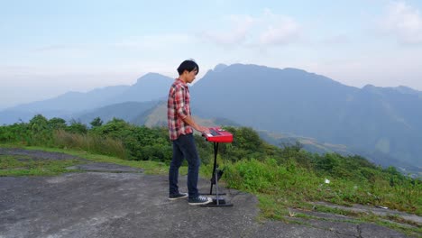 Eine-Stationäre-Aufnahme-Eines-Mannes-Von-Rechts,-Der-Auf-Seiner-Elektronischen-Musiktastatur-Spielt,-Mit-Blick-Auf-Die-Berge-Im-Hintergrund