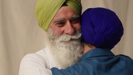 close up studio shot of senior sikh father embracing adult son both wearing turbans against plain background