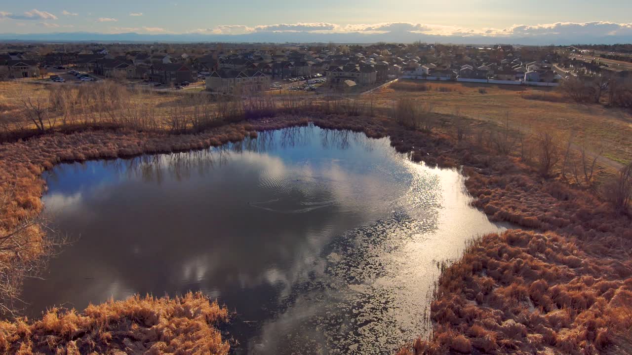 A drone backs up over a reflective pond revealing trees and clouds and the Rocky Mountains beyond. Filmed in Greeley Colorado