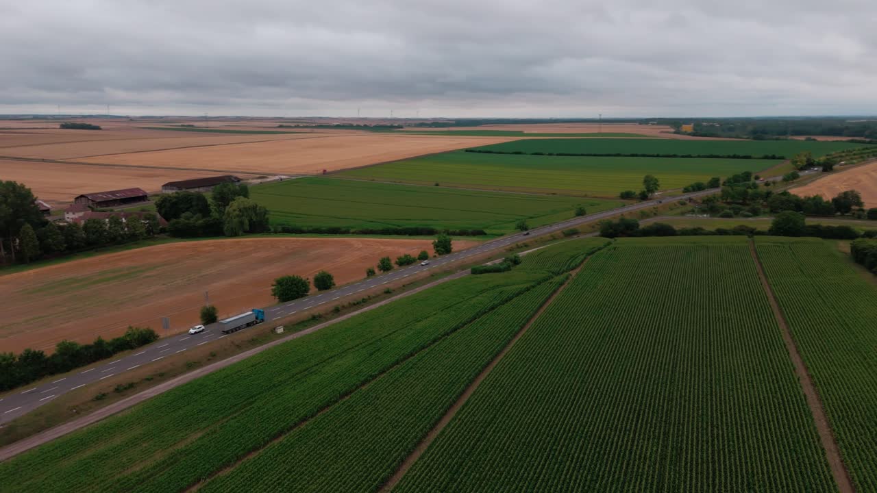 Aerial drone oftruck and cars driving on a road through the vast French countryside. A patchwork of green and brown agricultural fields stretches to the horizon under a cloudy sky