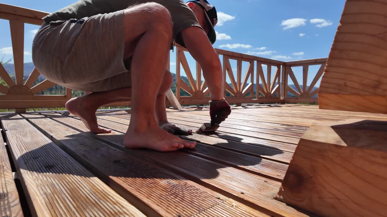 Man oils wooden deck boards with brush on a sunny summer day. Scenic mountain view, blue sky, and decorative railing highlight outdoor DIY wood protection and home improvement