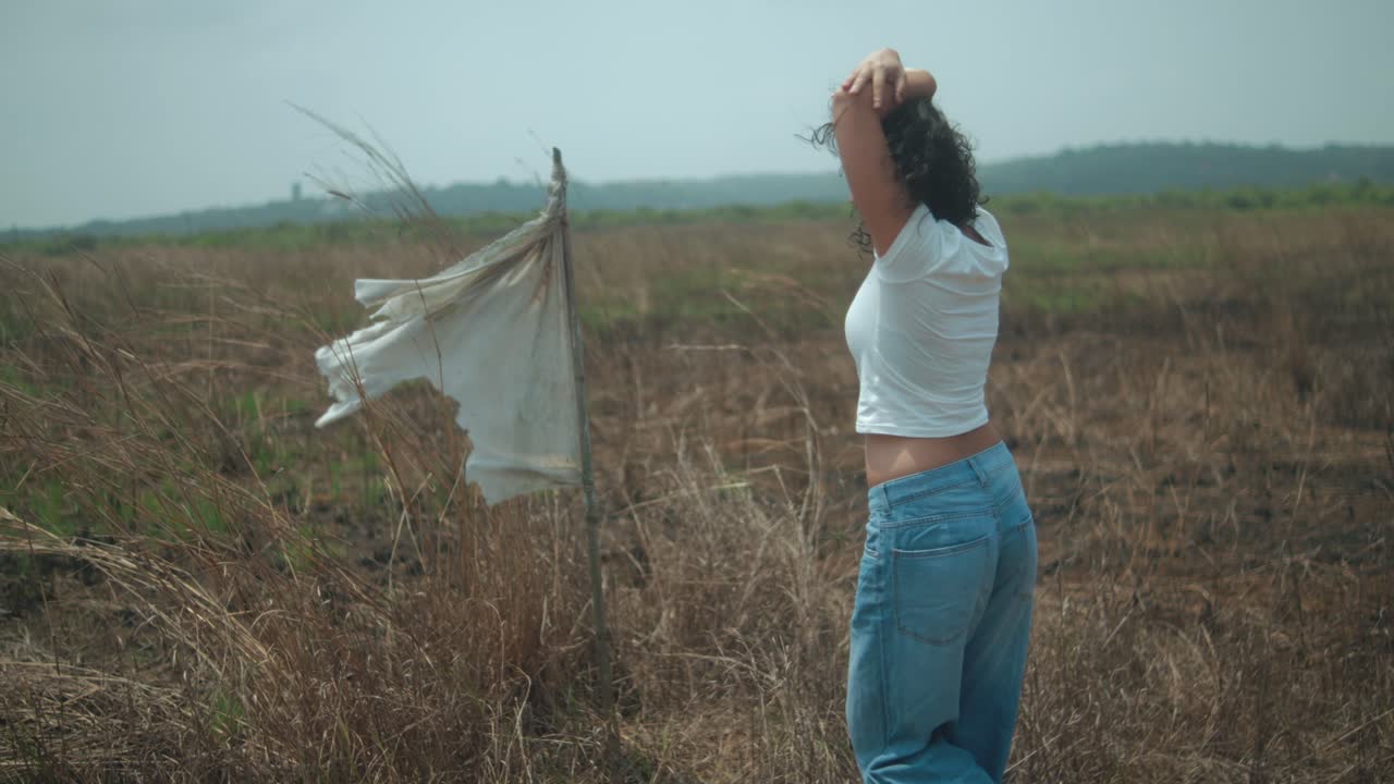 Young woman standing in a desolate dry grass field, raising her arms to the sky in a gesture of freedom, surrender, and emotional release, with a tattered white flag next to her