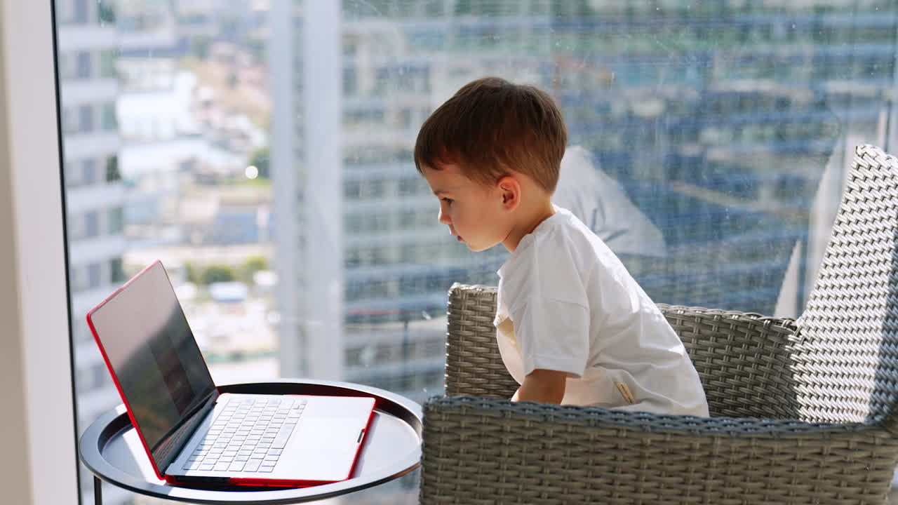 Boy using laptop on balcony in modern city. Child sitting on balcony chair using red laptop with city skyscrapers in background