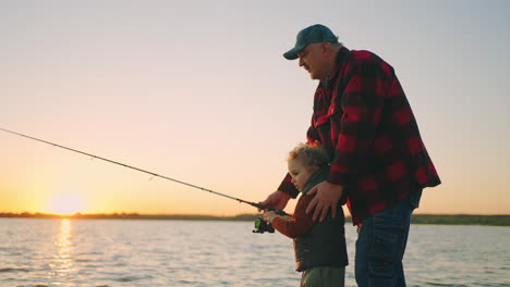 old fisher and little grandson are catching fish by rod in river or lake shore rest in nature in evening