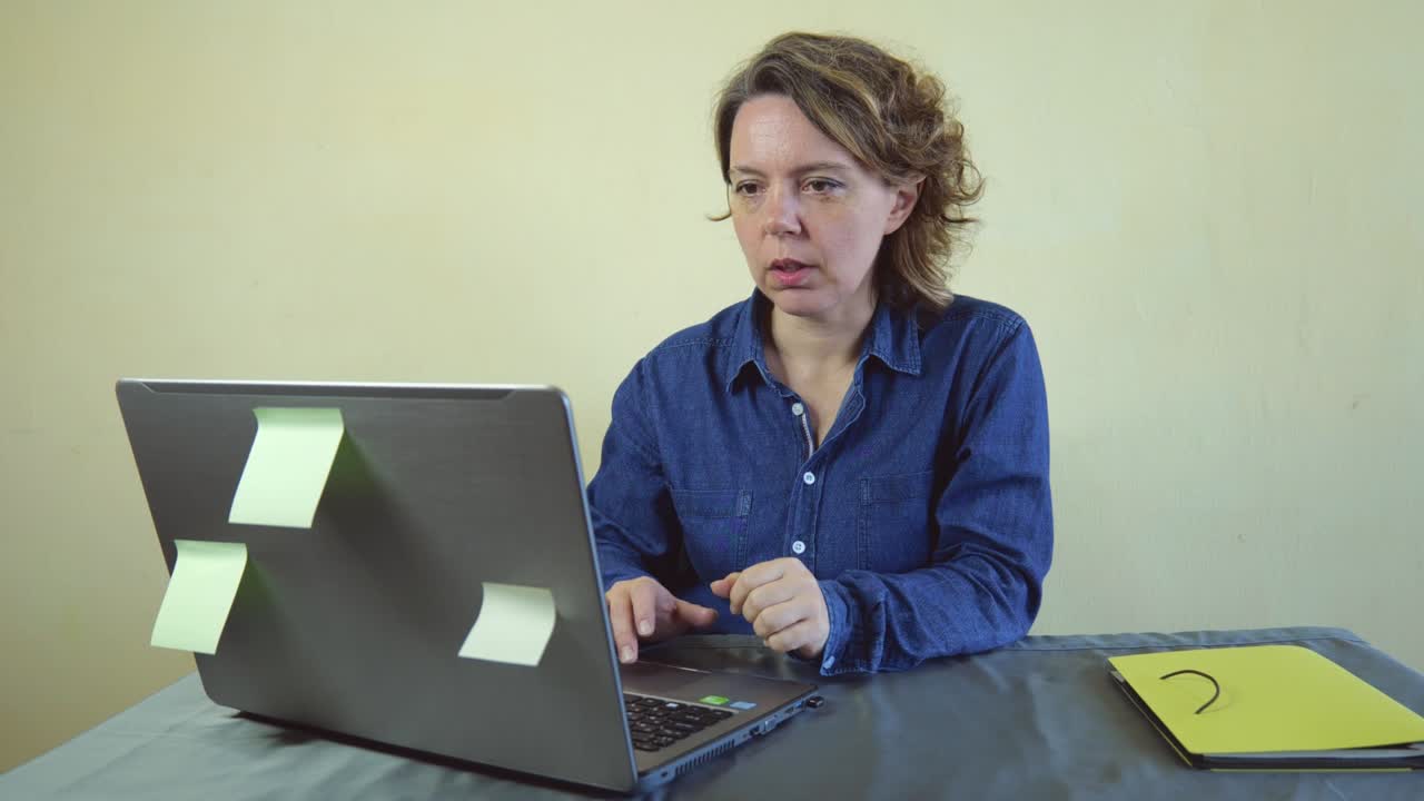 An adult forty-year-old woman in a blue shirt works at a laptop. Woman freelancer