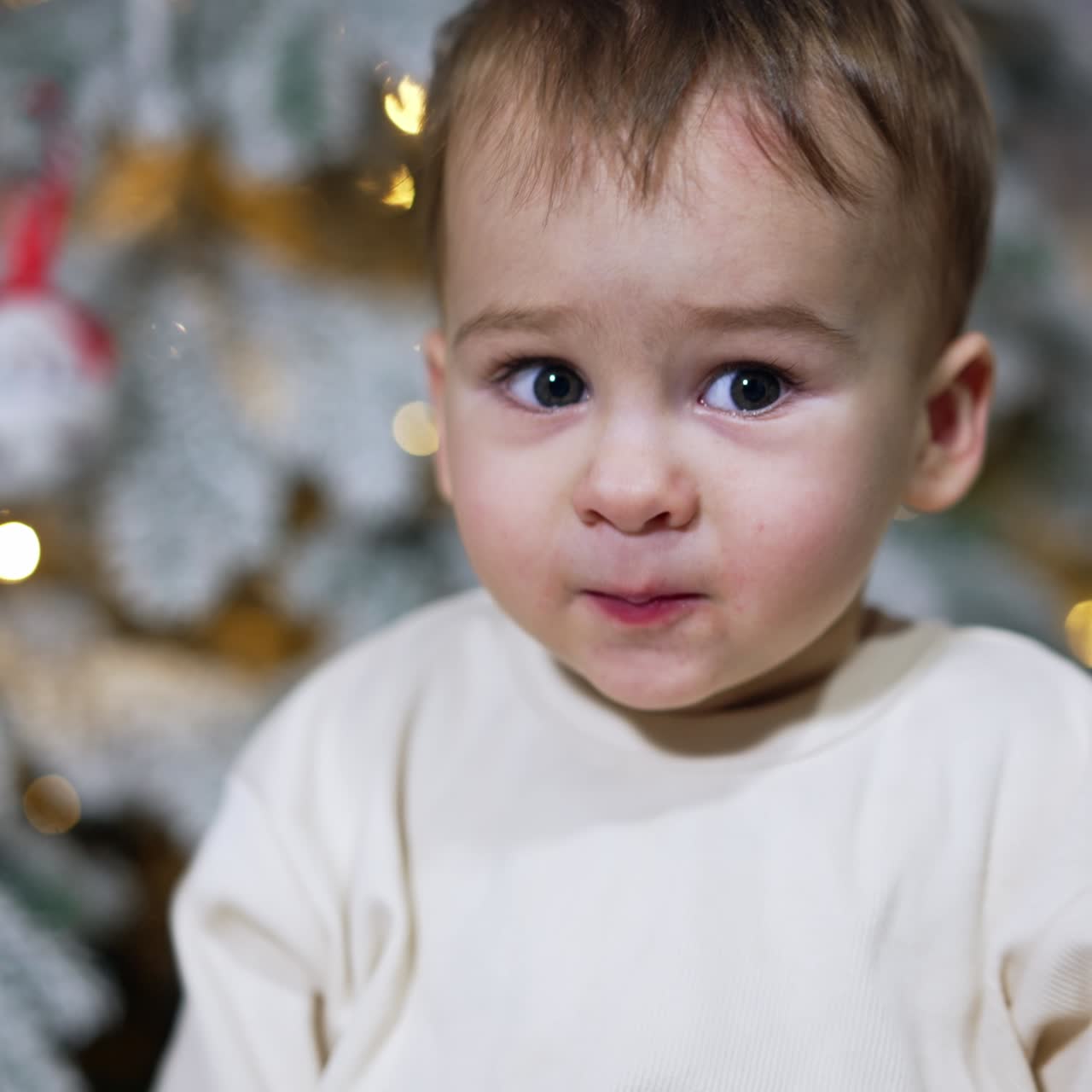 Lovely boy tries candy cane and waves hands excitedly. Child turns back to the Christmas tree behind