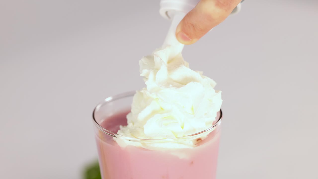 Hand dispenses whipped cream onto pink strawberry milkshake in glass, bright lighting, close-up view