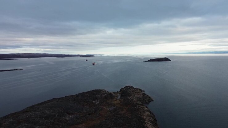 Elevated drone view tracking along rocky coastline toward open water horizon under overcast Arctic summer skies near shipping channels