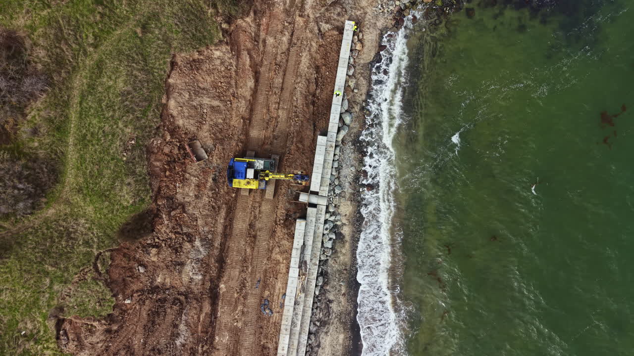 Cantiere per la costruzione di un molo lungo la riva con macchinari e operai presenti vicino all'acqua, di giorno.