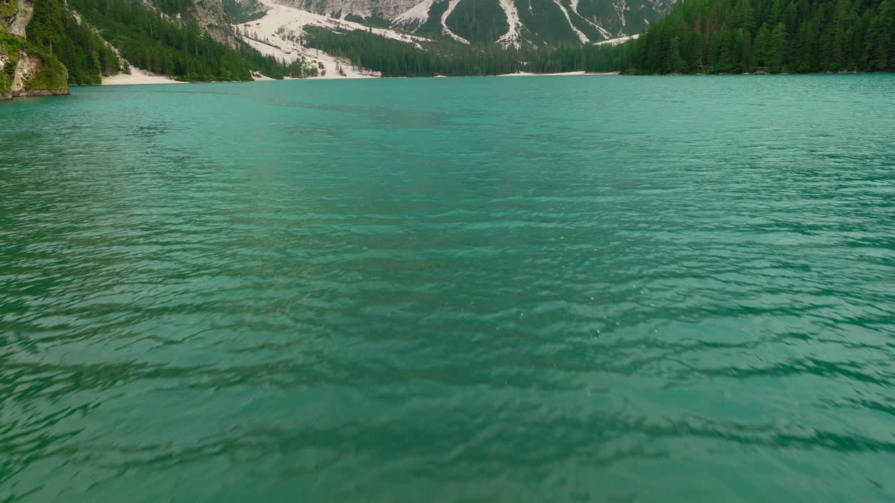 Low drone flight over the tranquil waters of Lake Braies on a cloudy spring day, revealing towering Dolomite peaks emerging from the misty mountain backdrop