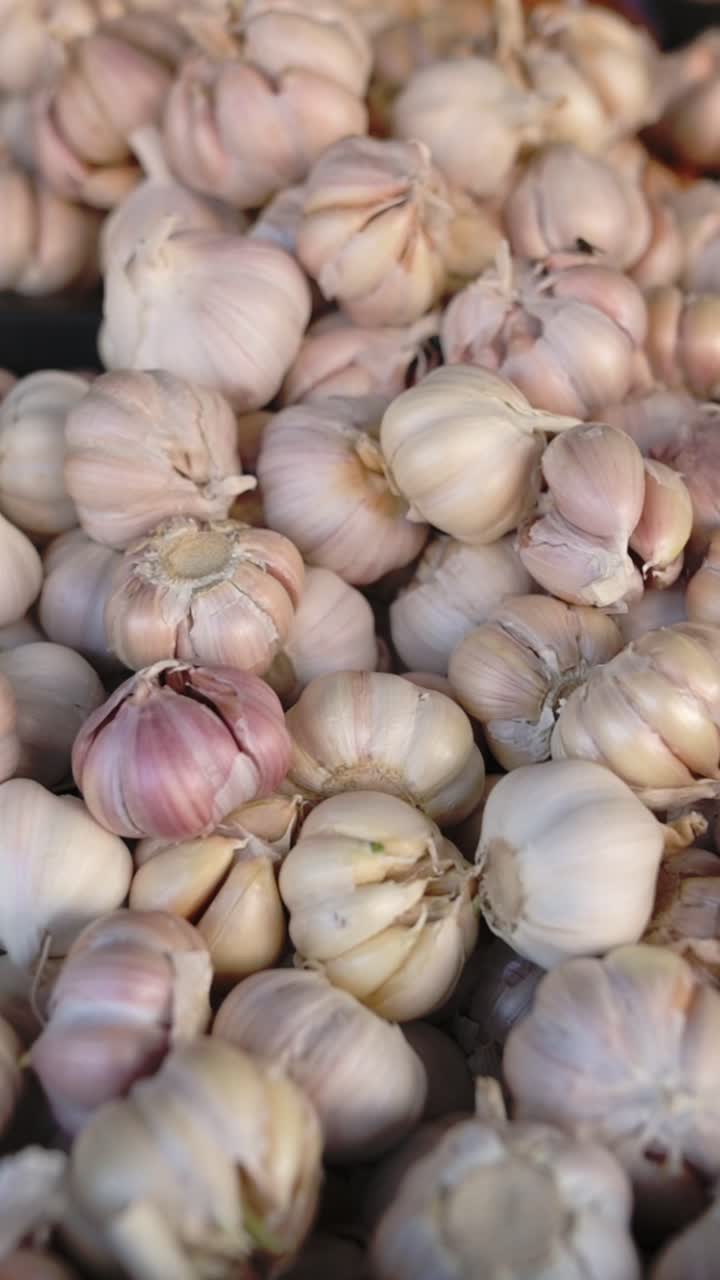Schermo verticale. Pila di aglio fresco al mercato di strada vietnamita. Primo piano di bulbi impilati con spicchi rosati e bucce cartacee, toni terrosi e illuminazione naturale. Prodotti pronti per la vendita che mettono in risalto consistenza e aroma.