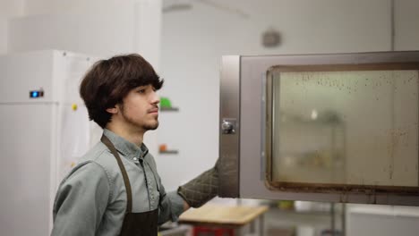 joven panadero sacando pasteles frescos del horno en la cocina de la panadería. vista lateral del chef masculino abriendo el horno y tomando bandeja con pasteles sabrosos cocinando en la cocina moderna