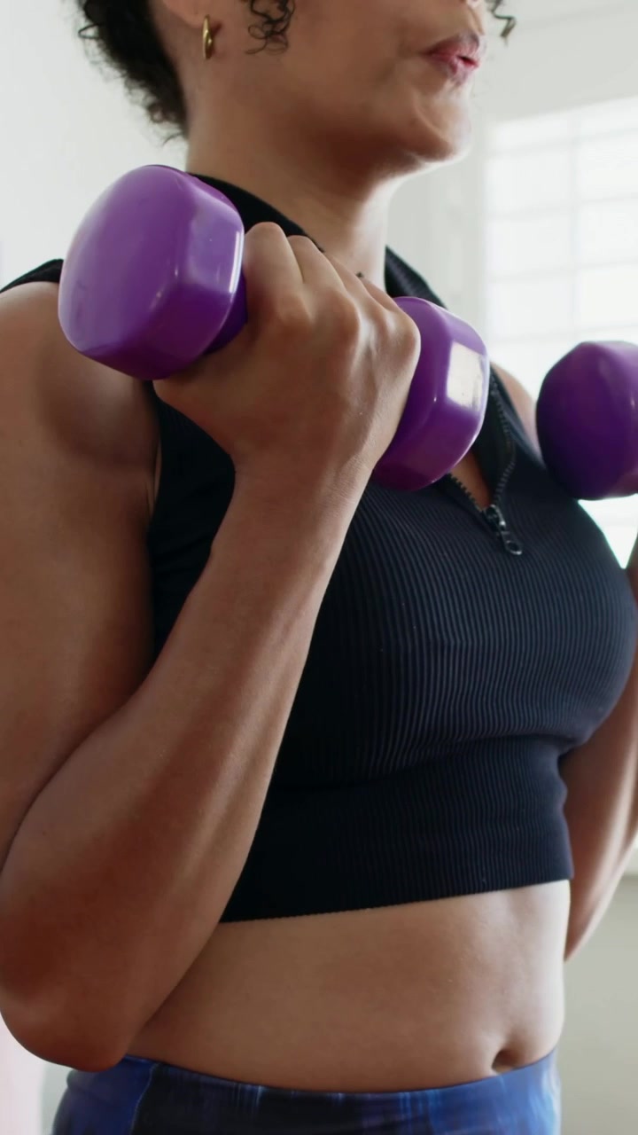 Vertical video: Starting from rest woman in cropped top curling purple weights at home to tone arms