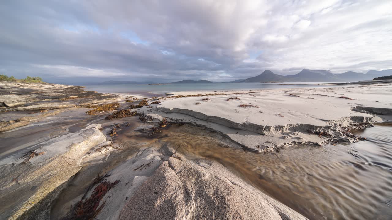 un lapso de tiempo de un arroyo poco profundo que fluye a través de la playa de arena como nubes girando por encima y las olas ruedan lentamente en la arena