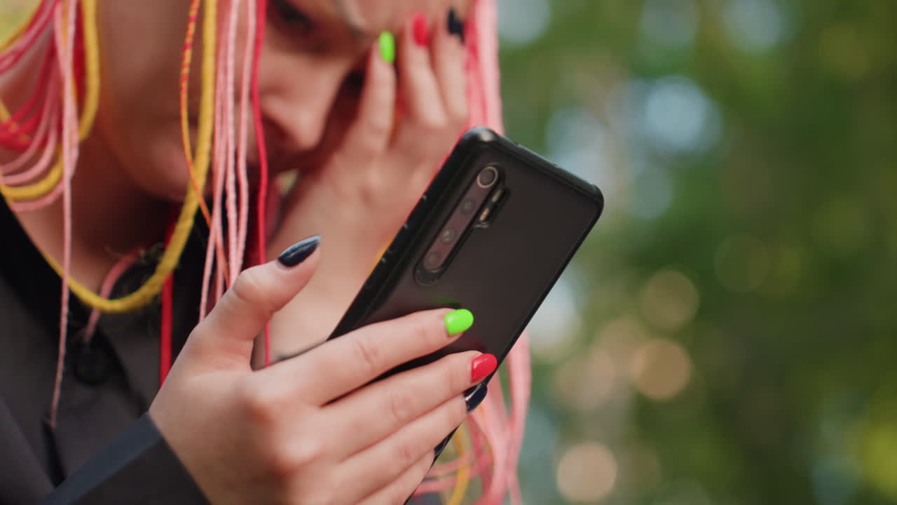 걱정스러운 표정으로 휴대폰을 쳐다보는 백인 여성, 얼굴 주변으로 다채로운 땋은 머리가 늘어져 있고, 네온 매니큐어가 화면을 두드리고 있으며, 흐릿한 녹색 배경의 벤치에서 긴장된 야외 분위기를 연출합니다.