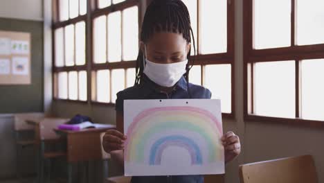 Girl-wearing-face-mask-holding-a-rainbow-painting-in-class-at-school-