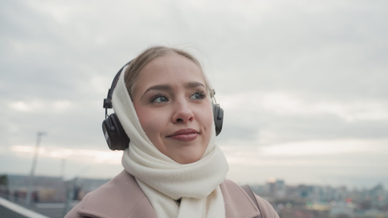 Donna che si gode l'aria aperta. Ragazza con le cuffie su un ponte. Donna caucasica sorridente con sciarpa e cuffie all'aperto. Giovane donna rilassata appoggiata al ponte con vista sulla città e cielo coperto.