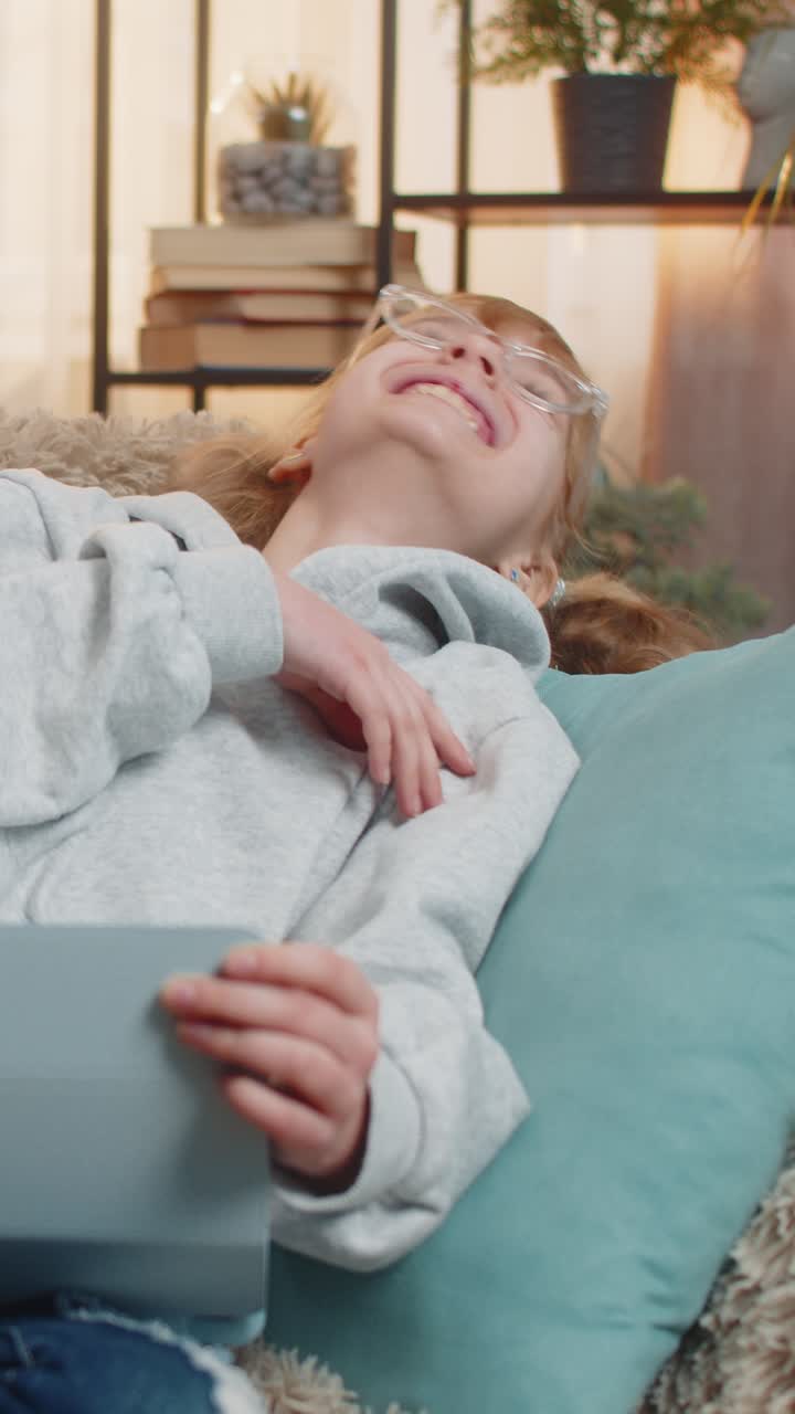 Young teenager girl enjoys watching online cartoons on tablet screen at home giggling laughing