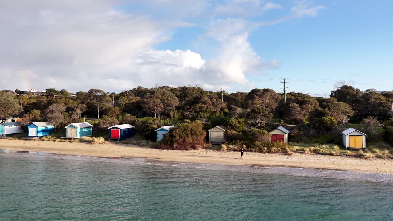 Drone pans over vibrant beach huts, sandy shoreline, and calm water in soft afternoon light