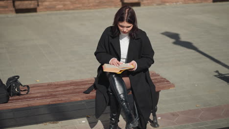 close-up of young woman reading a book on a sunny day, seated on a bench in an urban setting, her focus is on the book, with her hair and the background gently blurred