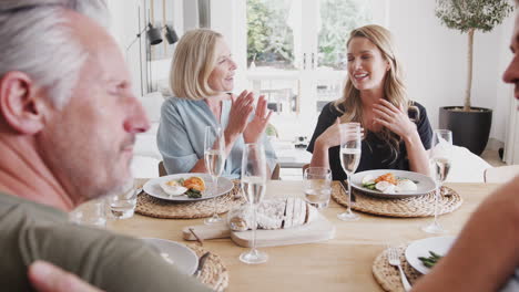 Family-With-Senior-Parents-And-Adult-Offspring-Sharing-Good-News-Before-Eating-Meal-Around-Table