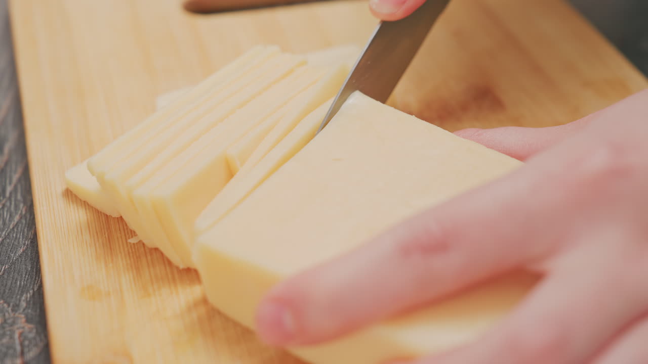 extreme close up of person slicing block butter into thin slices with knife on wooden cutting board during kitchen preparation, showing detailed texture of butter, knife blade