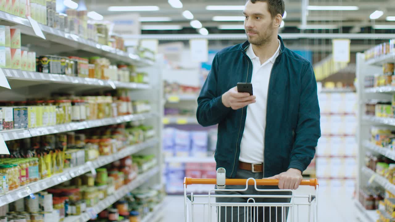 en el supermercado: un hombre guapo revisa un estante con productos enlatados, coloca una lata en su carrito de compras y continúa con su lista de compras caminando por diferentes secciones.