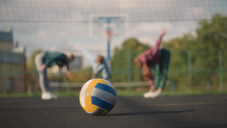 close-up of volleyball resting on outdoor court with blurred view of athletes performing stretches and bends in the background, sports facility, and building visible