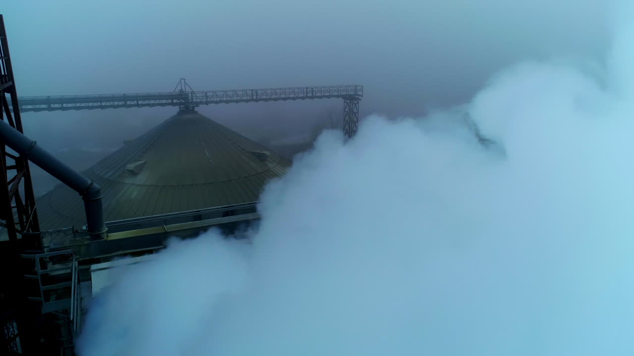 A number of pipes producing thick white smoke at agricultural plant. Rounded roof of silo tank at backdrop. Grey foggy day background.