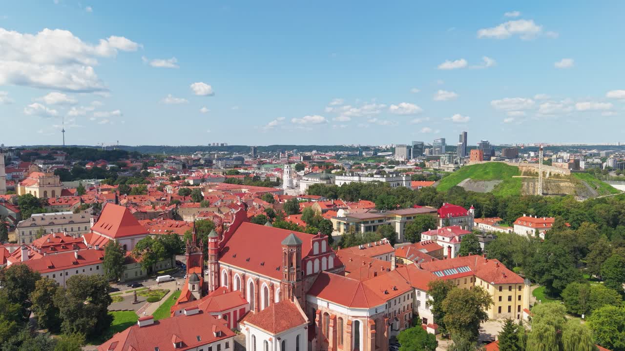 Aerial panoramic view of Vilnius Old Town, featuring red rooftops, baroque churches, and Gediminas Hill with modern cityscape in the background on a sunny summer day