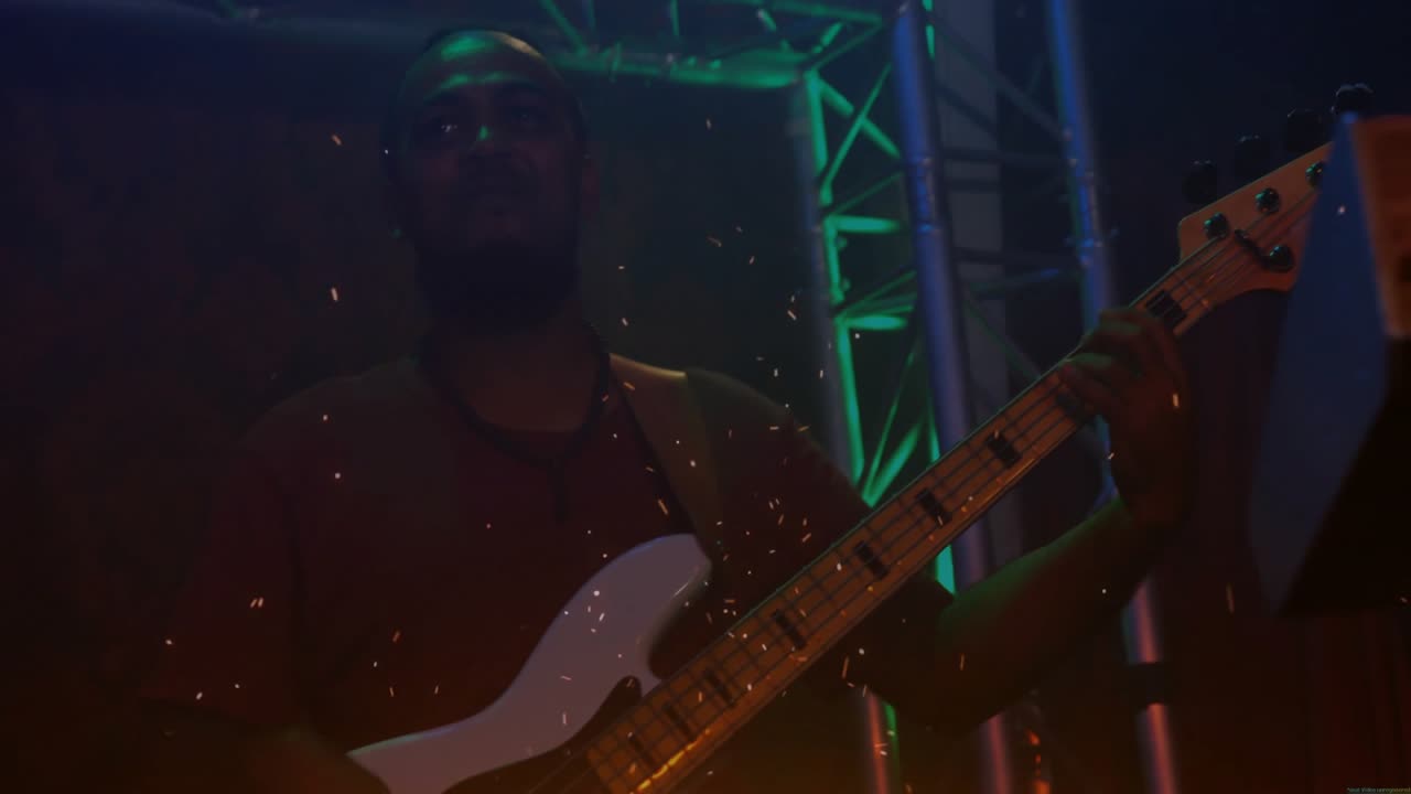 Männlicher Musiker spielt Bassgitarre auf schwach beleuchteter Musikbühne, mit schwebenden Funken und LED-Traversenanimation.