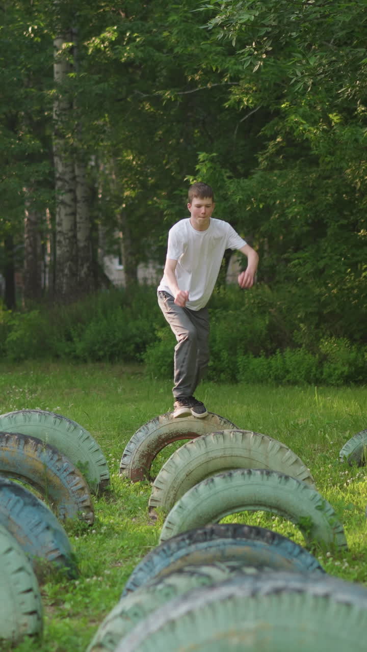 Front view of a young boy in a white shirt and gray pants, jumping on a row of tires in a grassy field, captured in slow motion with green trees in the background