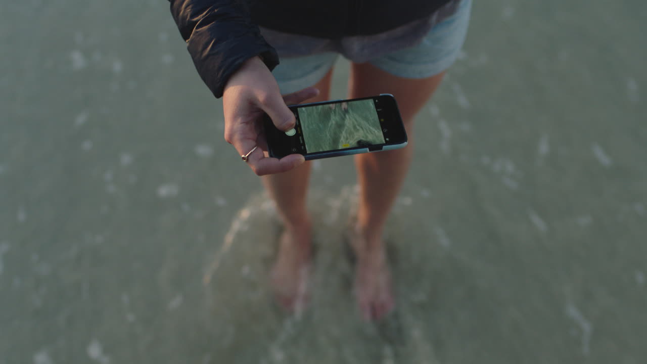 mujer en la playa tomando una foto de las olas que salpican suavemente los pies usando tecnología de cámara de teléfono inteligente compartiendo experiencia de viaje
