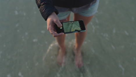 mujer en la playa tomando una foto de las olas que salpican suavemente los pies usando tecnología de cámara de teléfono inteligente compartiendo experiencia de viaje