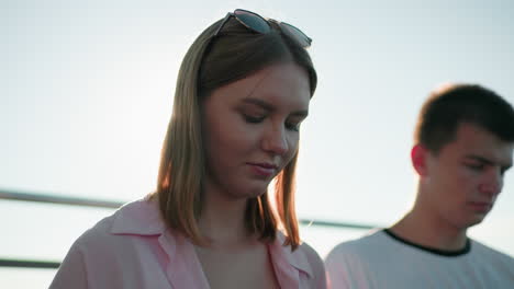 close-up of young woman with glasses on head, sun backlight creating halo, and focused young man beside her, against a railing and sky backdrop