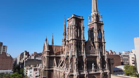 Aerial-pan-shot-capturing-details-facade-of-gothic-style-church-of-capuchinos,-Sacred-Heart-of-Jesus-with-birds-flying-pass-the-building-against-clear-blue-sky-in-downtown-Cordoba-city,-Argentina