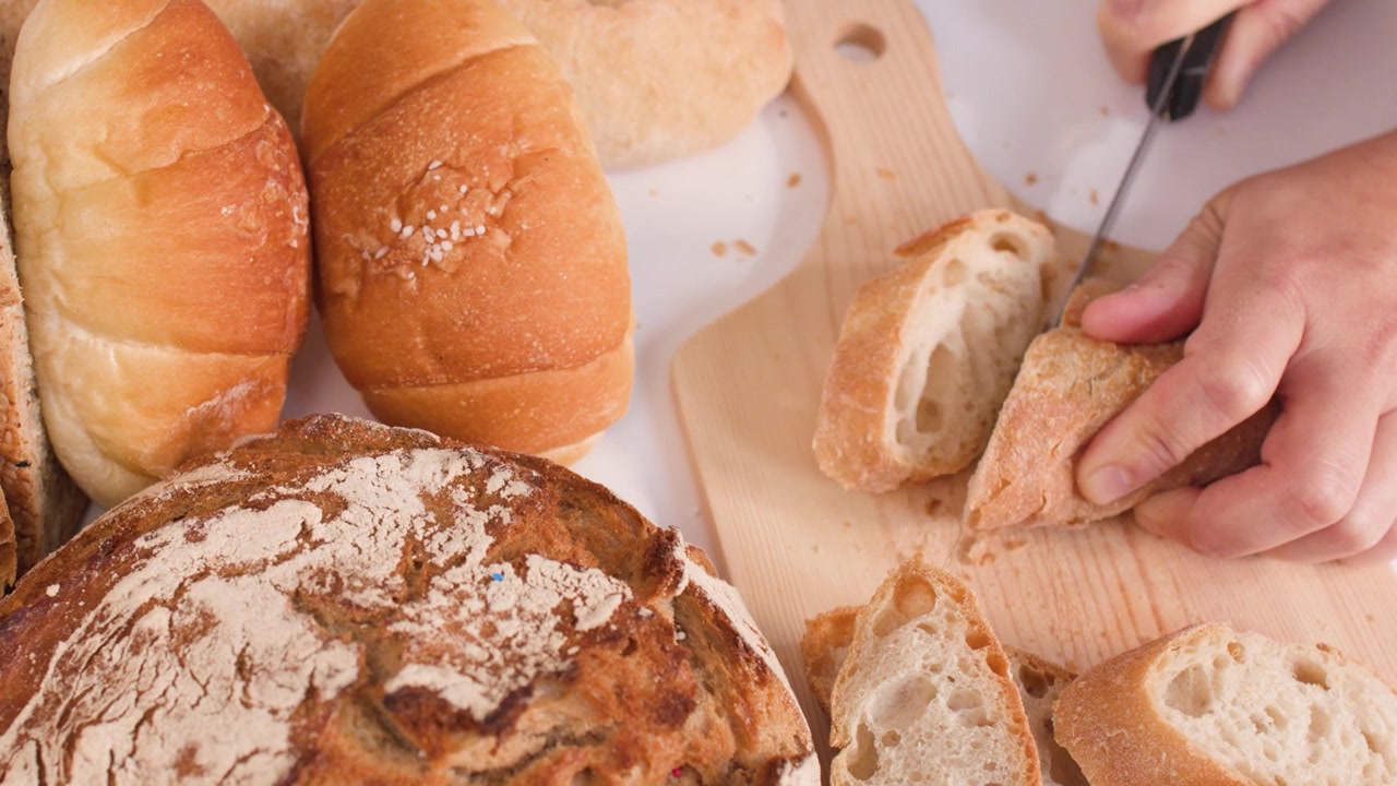 Close-up of hands slicing crusty bread on a wooden board amidst various bakery items.