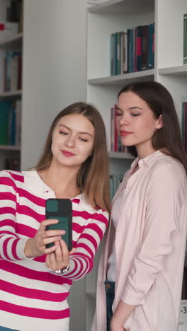 girl friends take selfie together near bookshelves. happy students shoot blog about college life for social media in library. women bloggers in bookstore