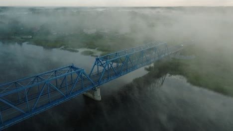 Vista-Aérea-Del-Puente-De-Acero-Sobre-El-Río-Lielupe-En-Una-Mañana-Soleada-De-Verano,-Niebla-Elevándose-Sobre-El-Río,-Autos-Conduciendo,-Amplio-Disparo-Ascendente-De-Drones,-Inclinación-Hacia-Abajo