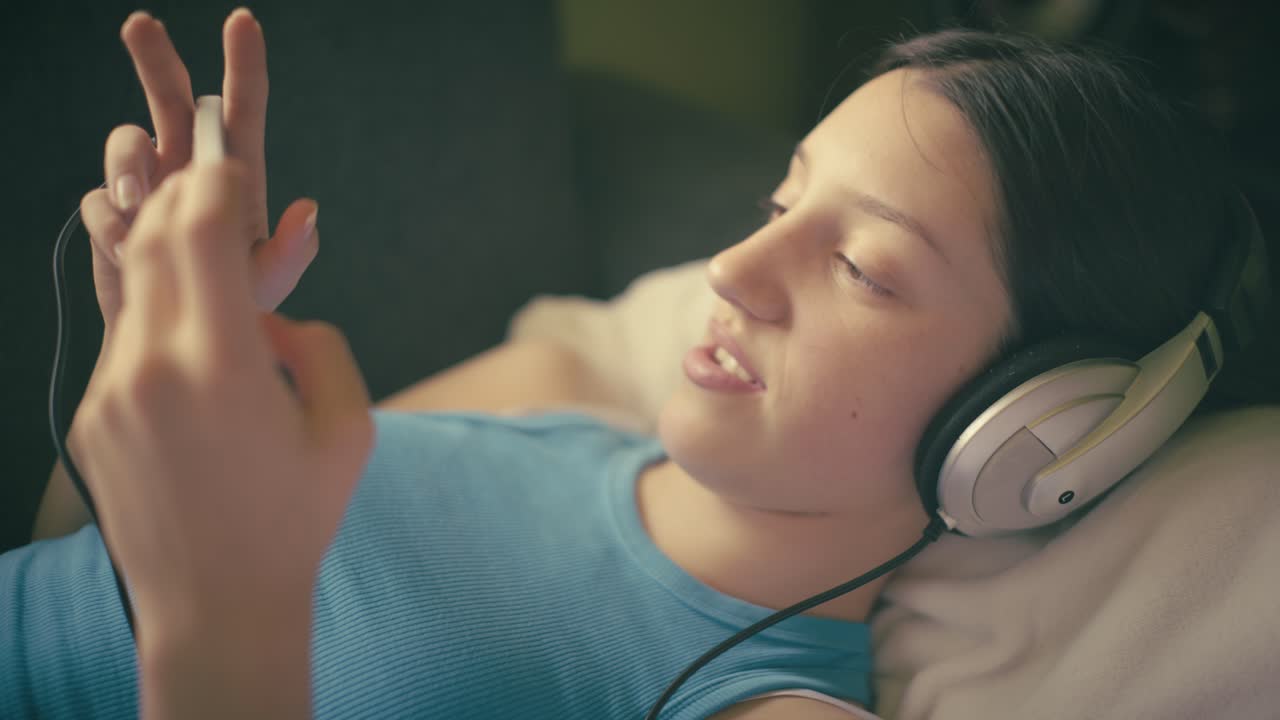 Teen girl relaxing at home, listening to music with headphones and using her smartphone. A calm, intimate moment of digital leisure in a cozy setting.