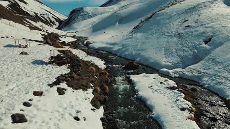 Es-Entfaltet-Sich-Eine-Malerische-Winterszene-Mit-Einem-Kristallklaren-Fluss,-Steinigen-Ufern-Und-Einer-Rustikalen-Brücke,-Die-Die-Ruhe-Der-Natur-Inmitten-Der-Schönheit-Der-Berge-In-Einklang-Bringt