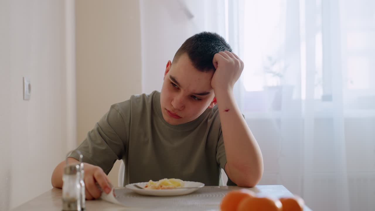 Unhappy boy sits at dining table with plate of food untouched, resting head on hand with sad expression, natural daylight through curtain capturing moment of refusal and frustration